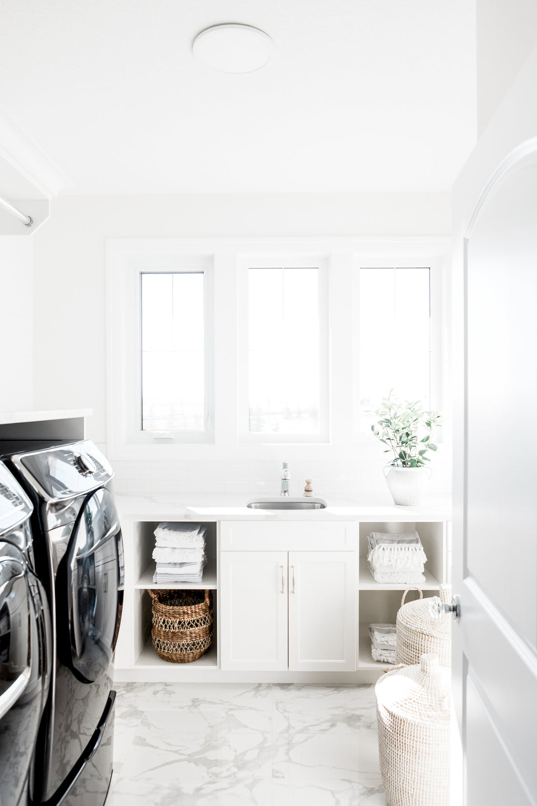 Bright white laundry room with front-loading washing machine and dryer on a pedestal, folded white towels, two woven laundry baskets.